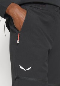 Black athletic pants featuring an orange zipper pull, side pocket, and a white logo on the thigh. The fabric has a smooth texture.