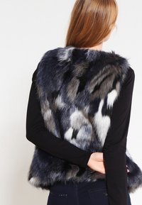 Faux fur-väst i nyanser av blått, grått och vitt med en plyschig textur. Har inga knäppningar och en rundad halsringning, bärs över en svart topp.
