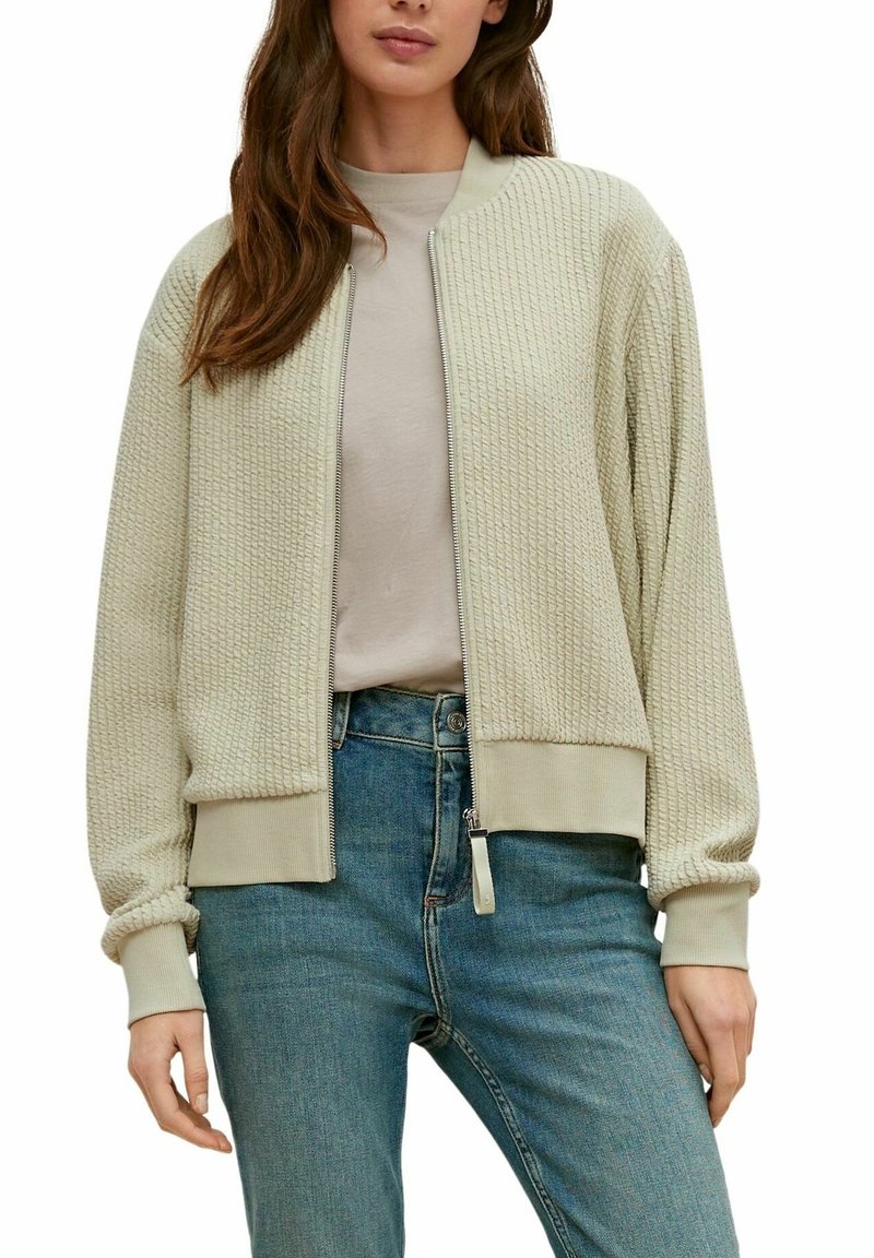 Femme portant une veste bomber zippée beige clair texturée sur un haut beige et un jean bleu, debout, les mains détendues le long du corps.