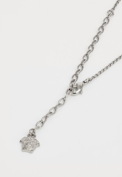 Collier en argent avec maillons ajustables et un pendentif en forme de tête de Méduse près du fermoir en mousqueton sur un fond blanc.