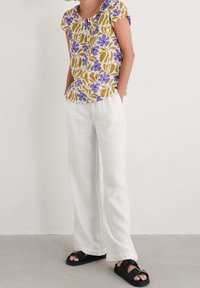 Top con motivo floreale giallo e viola, abbinato a pantaloni larghi in lino bianchi e sandali neri a scivolo. Outfit casual con una vestibilità rilassata.
