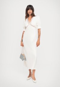 Witte jurk tot de knie met pofmouwen en kantdetails bij de halslijn, gecombineerd met een lichtgrijze handtas en beige puntige schoenen.
