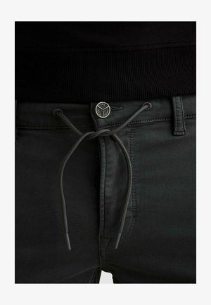 Gros plan sur un pantalon noir avec un cordon de serrage passé à travers des œillets et des coutures cousues, associé à un vêtement noir à taille côtes.