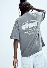 Graues, oversized T-Shirt mit einem weißen Logo "stwd" und Text auf dem Rücken. Der Stoff ist weich und hat eine lässige, lockere Passform. Getragen mit hellblauen Jeansshorts.