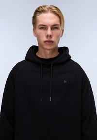 Felpa nera con vestibilità rilassata, dotata di cappuccio con cordino, polsini a costine e un piccolo logo sul petto. Tessuto morbido, design minimale.