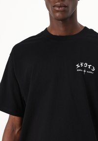 Maglietta nera in cotone con maniche corte, con testo e logo bianchi sul lato sinistro del petto. Design semplice e classico a girocollo.