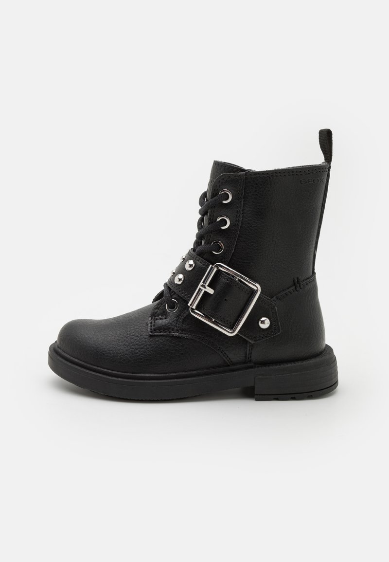 Bottine en cuir noir avec lacets, une large sangle à boucle argentée et des clous en métal, posée sur une surface blanche.