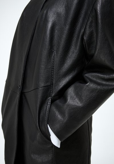 Nahaufnahme eines Ärmels einer schwarzen Lederjacke, in dessen Seitentasche teilweise eine Hand steckt, vor einfachem Hintergrund.