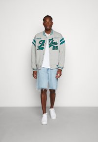 Gray varsity jacket with teal accents, front buttons, and graphic lettering, paired with light blue denim shorts and white sneakers.