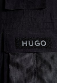 Czarna tkanina z naszytą kieszenią i rzepem z naszywką z napisem "HUGO" w szarych wielkich literach.