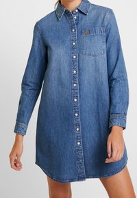 Denimklänning i medelblå nyans, med knappstängning fram, lång ärm, en bröstficka och en rundad nederkant.