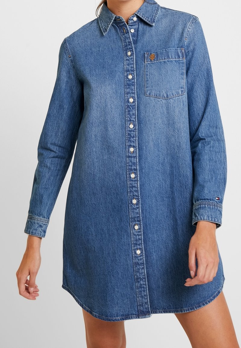 Denimklänning i medelblå nyans, med knappstängning fram, lång ärm, en bröstficka och en rundad nederkant.