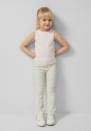 Young girl with blonde hair wearing a sleeveless pink top, floral pants, and white sneakers standing against a gray wall with hands on hips.