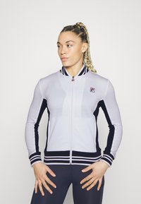 Hvid atletisk zip-jakke med marineblå sidelapper, stribet krave og Fila-logo. Har tætsiddende design og ribbede manchetter.