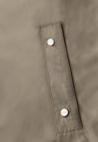 Gros plan sur un tissu beige montrant une poche verticale avec deux boutons-pression argentés sur le rabat.