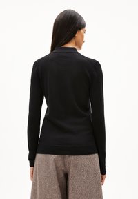 Frau mit langen dunklen Haaren, die ein schwarzes Langarmoberteil und lockere grau-braune Hosen trägt, mit dem Rücken zu einer weißen Hintergrund.