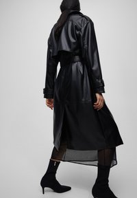 Femme portant un trench en cuir noir ceinturé, une jupe noire transparente et des bottines noires à talons, se tenant devant un fond gris uni.