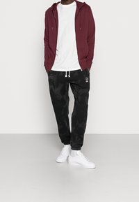 Maroon zip-up hoodie, white t-shirt, black tie-dye joggers with embroidered accents, and white sneakers. Casual, comfortable fit.