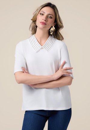 Blouse blanche avec un col structuré orné de perles horizontales, manches courtes et coupe décontractée, associée à un pantalon en jean bleu.