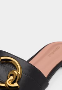 Μαύρο δερμάτινο σανδάλι slide με μεγάλο χρυσό κυκλικό αγκράφα και ροζ ανοιχτό πέλμα, μερική πλευρική όψη σε λευκό φόντο.