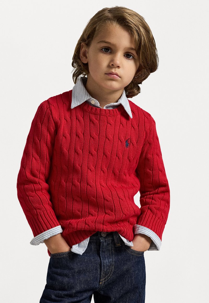 Roter, kabelgestrickter Pullover mit Rundhalsausschnitt, über einem hellblauen gestreiften Hemd getragen. Dunkelblaue Denim-Jeans mit klassischer Passform.