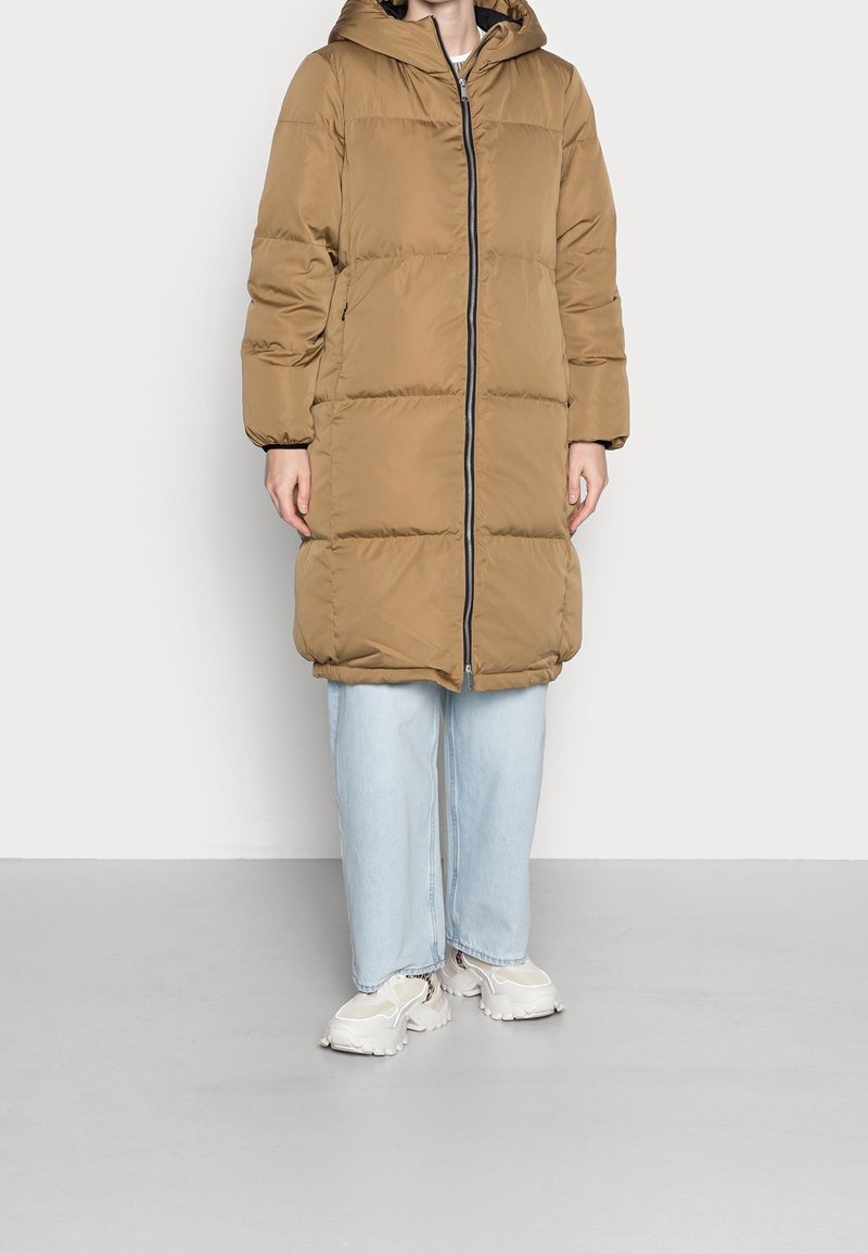 Cappotto puffer marrone con cappuccio, dotato di zip a tutta lunghezza e polsini elasticizzati. Indossato sopra jeans a gamba larga azzurri chiari e sneaker bianche.