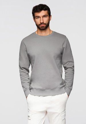 Mężczyzna o ciemnych, kręconych włosach i brodzie, ubrany w szarą bluzę i białe spodnie typu cargo, stojący z rękami w kieszeniach na jednolitym tle.