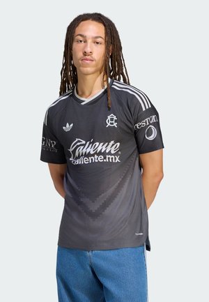 Junge Person mit langen Dreadlocks steht, trägt ein schwarzes Adidas-Trikot mit weißen Logos und blaue Jeans vor einem einfarbigen Hintergrund.