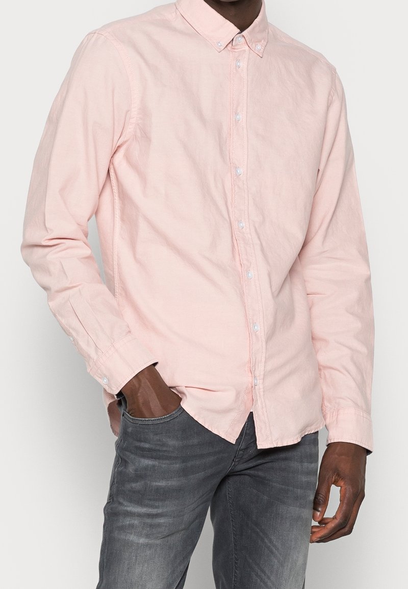 Homme portant une chemise rose clair à boutons avec col et un jean gris foncé, une main dans la poche, sur un fond uni.