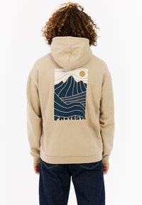 Beige hoodie met een grafische print van gestileerde bergen en golven in marineblauw en crème, met de tekst "PROTEST" onderaan. Gemaakt van een katoenmix.