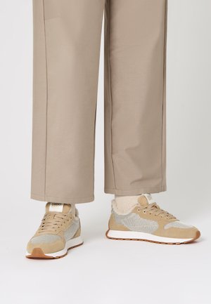 Sneaker beige con texture in rete e suede, dotate di un colletto imbottito, suola intermedia bianca e suola in gomma. Indossate con calzini e pantaloni chiari.