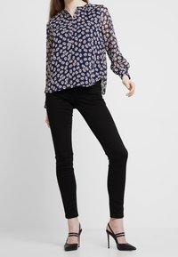 Marinblus med blommönster, genomskinliga långa ärmar och krage; kombinerad med svarta slim-fit byxor och svarta klackskor med remmar.