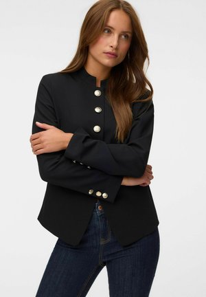 Vrouw met lang bruin haar die een zwart gebuttonde blazer en donkerblauwe jeans draagt, armen gekruist, kijkt iets opzij tegen een effen achtergrond.
