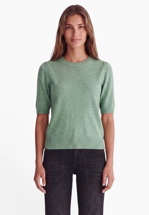 Femme aux longs cheveux bruns portant un haut tricoté vert à manches courtes et un pantalon noir, debout devant un fond blanc.