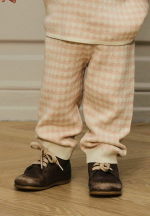 Bambino con pantaloni check beige e scarpe marroni scure con lacci, in piedi su un pavimento in legno contro uno sfondo di parete beige.