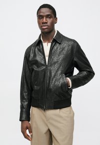 Μαύρο δερμάτινο μπουφάν τύπου bomber με ανάγλυφα κυματιστά σχέδια, κλείσιμο με φερμουάρ, ριμπ μανσέτες και τελείωμα, φορεμένο πάνω από μπεζ πουλόβερ και παντελόνι.