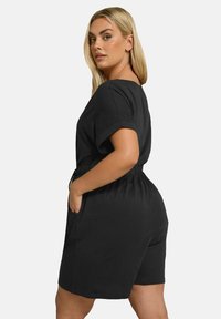 Svart bomullsbyxdress med korta ärmar, avslappnad passform, elastisk midja och sidofickor. Slät textur, enkel design utan synliga mönster.