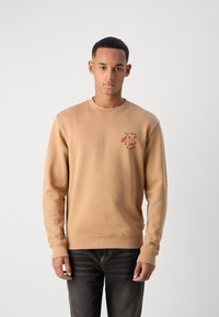 Beige sweatshirt made of cotton, featuring long sleeves and a round neckline. A small red logo is printed on the left chest.