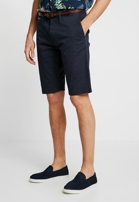Shorts en coton bleu marine à l'avant plat, poches latérales et coupe slim, assortis à des chaussures navy sans lacets et une chemise à motifs.
