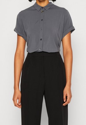 Osoba ubrana w szarą koszulę na guziki z krótkim rękawem, kołnierzykiem i podwiniętymi mankietami, włożoną do czarnych spodni z wysokim stanem.