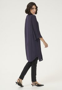 Haut léger à manches longues violet avec un design fluide, associé à un pantalon noir ajusté et des chaussures à talons ouverts noires.