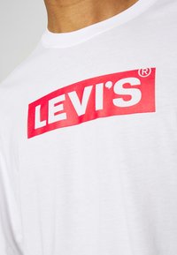T-shirt bianca in cotone con un grande logo rosso Levi's sul petto. Design a collo rotondo con texture liscia e maniche corte.