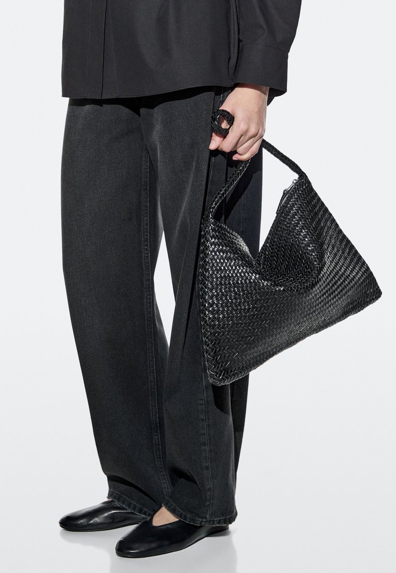 Personne tenant un grand sac à main en cuir tressé noir, portant un pantalon ample noir, des chaussures noires et un haut à manches longues noir sur fond blanc.