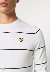 Maglione a righe grigio chiaro con strisce orizzontali nere e logo ricamato giallo sul petto sinistro. Texture lavorata a maglia, scollo tondo.