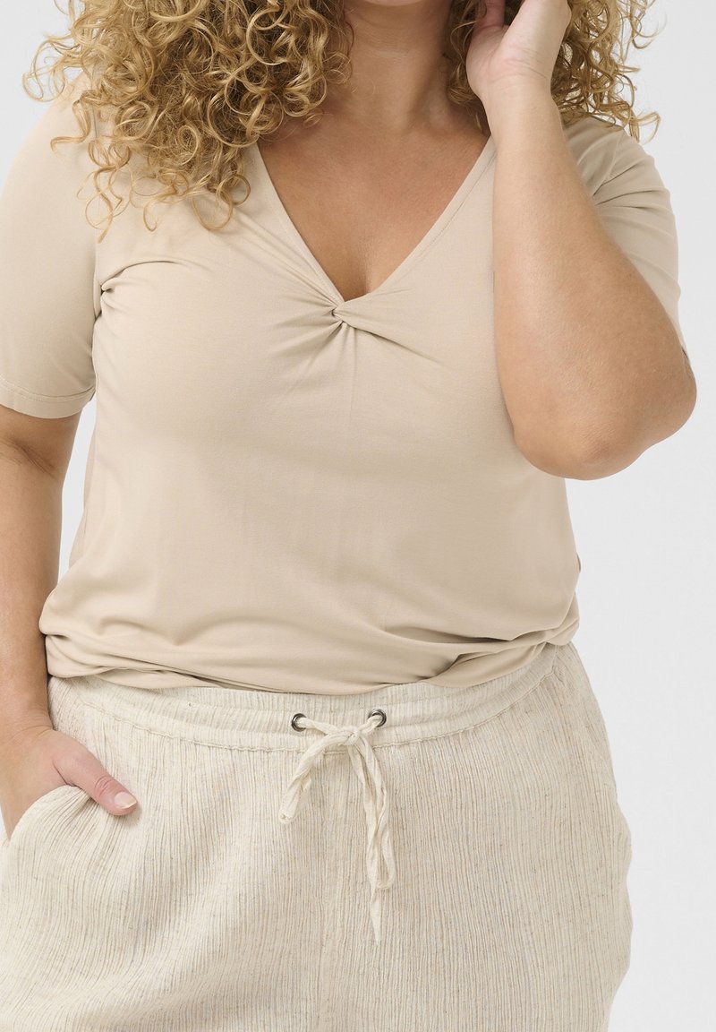 Femme aux cheveux bouclés portant un t-shirt beige à col en V avec manches courtes et un pantalon beige clair à cordon, une main dans la poche.
