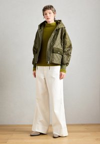 Olivenfarbiger, gesteppten Jacke mit Kapuze und Fronttasche, kombiniert mit einem gerippten Olivpullover und weißen, weit geschnittenen Hosen. Glatte graue Schuhe.