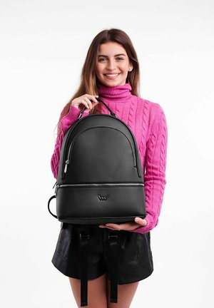 Černý batoh s kulatým tvarem, hladkým koženým povrchem, přední kapsou na zip a logem. Model má na sobě růžový svetr s copánkovým vzorem.
