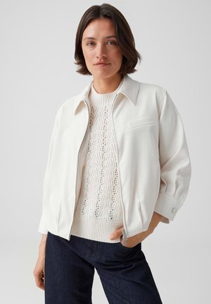 Frau mit kurzem dunklem Haar trägt einen weißen strukturierten Strickpullover unter einer weißen Reißverschlussjacke und dunkelblaue Hosen vor einem einfarbigen Hintergrund.