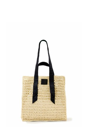 Sac fourre-tout en paille tissée beige avec de longues poignées en tissu noir et une petite étiquette carrée noire sur le devant.