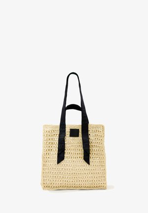 Bolso tote de paja beige trenzada con asas largas de tela negra y un pequeño parche cuadrado negro en el frente.